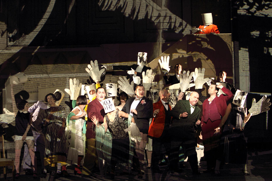 Actors on stage suits holding up big paper hands in ENO's Lulu