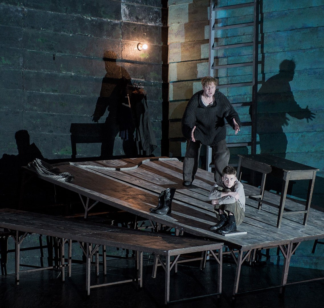 Man on stage stood menacingly behind a small boy who is crouched down