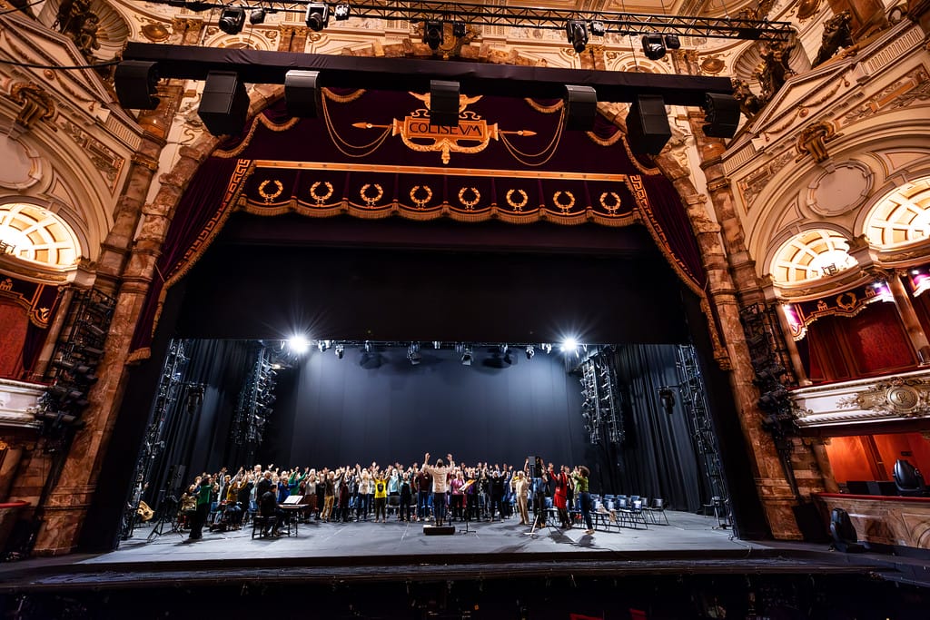 Various community groups attending an onstage singing workshop at the Coliseum. They are seated on the stage as a workshop leader talks to them.